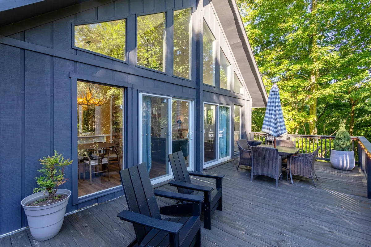 A serene exterior shot featuring a furnished deck with comfortable seating and expansive views of lush foliage and the surrounding landscape. The large windows connect the deck to the interior, reinforcing the seamless flow between indoor and outdoor living spaces.