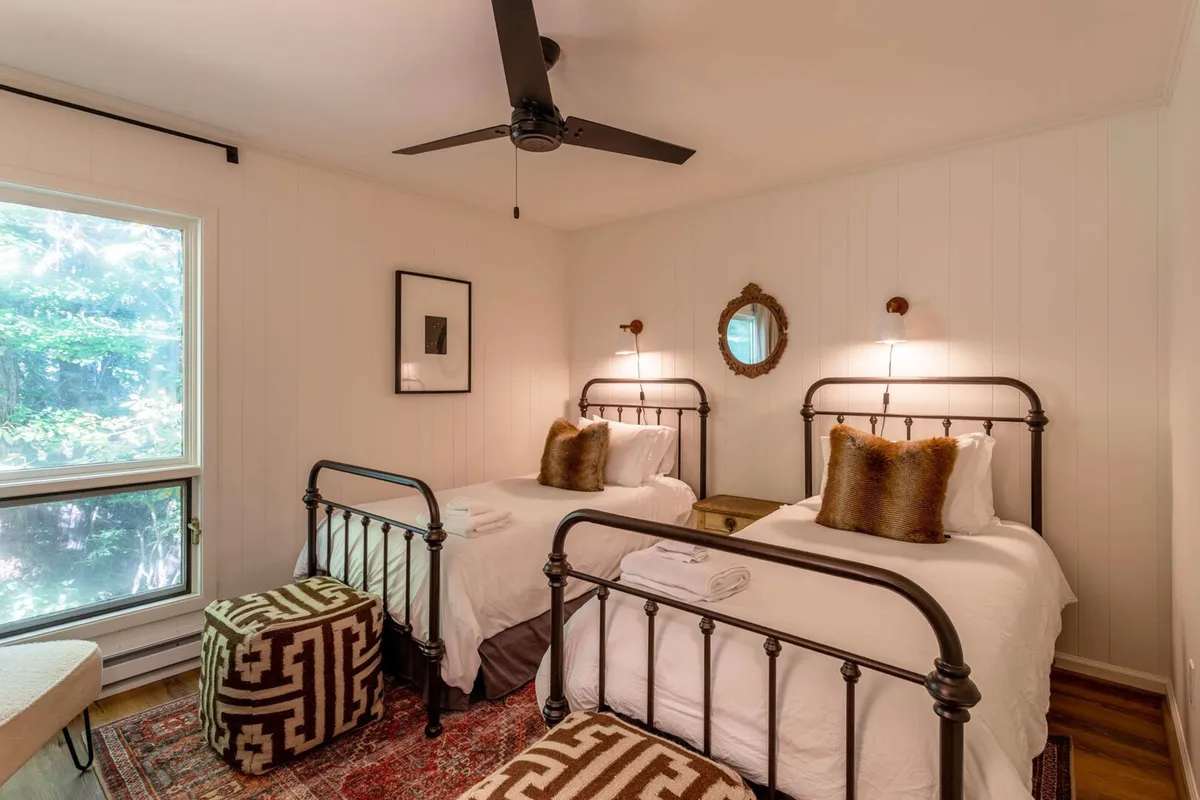 Two metal framed twin beds with decorative pillows are featured in a cozy, sunlit bedroom. Framed artwork and a round mirror add to the aesthetic appeal, while the window allows natural light to brighten the space, perfect for families or friends.
