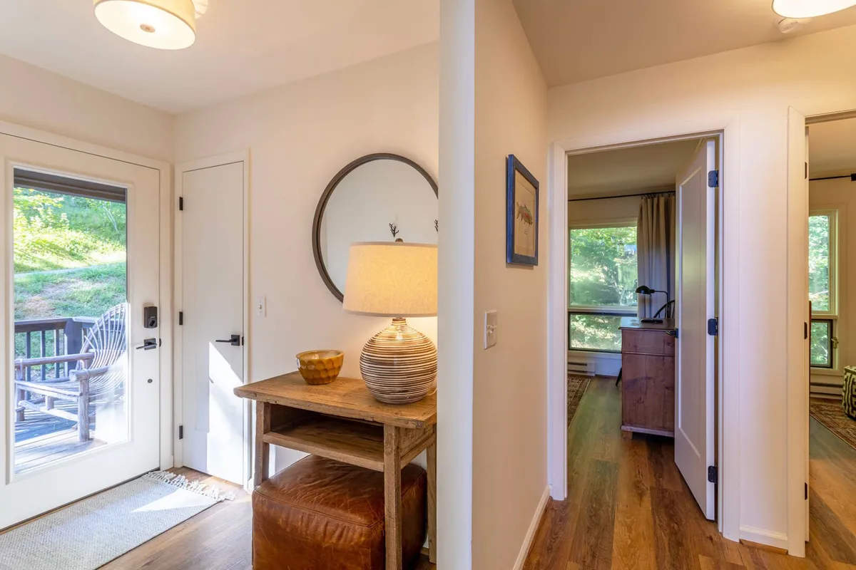 This image captures the entryway of Spruce House, featuring a welcoming table lamp, a decorative mirror, and tasteful artwork. Morning light streams in through the windows, creating a bright, inviting first impression that suggests arrival at a peaceful mountain retreat.