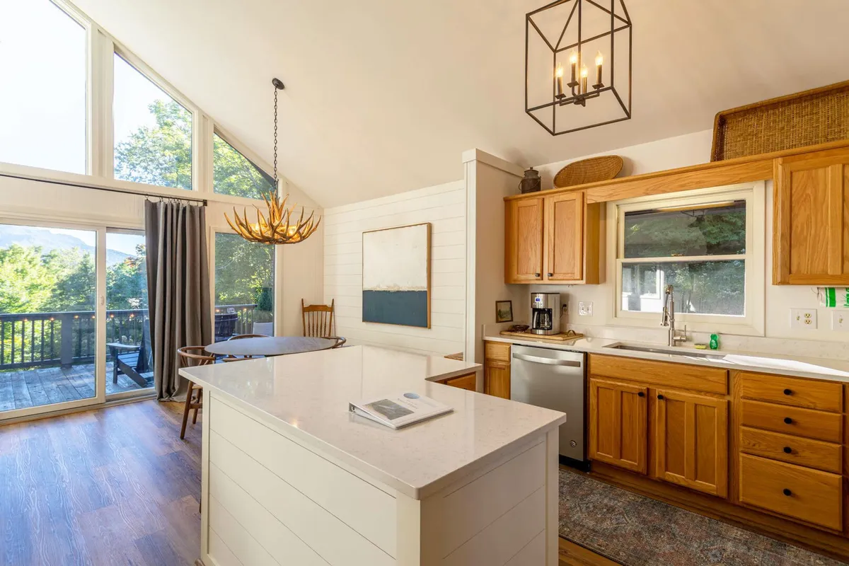 The view focuses on an inviting kitchen island, elegant chandelier, expansive windows, and wooden cabinetry. Morning light streams in, creating a warm, communal atmosphere for both cooking and sharing meals. The setup encourages both socializing and practical meal preparation.