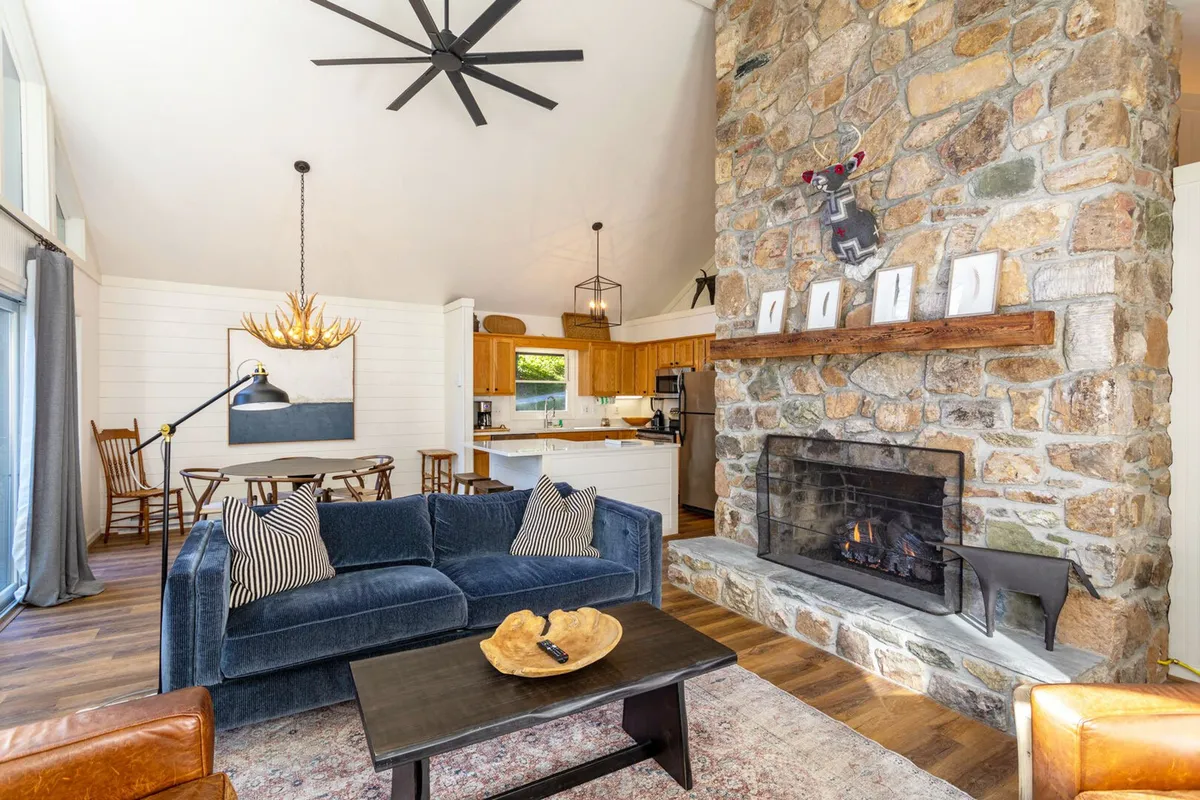 This image captures a warm and welcoming interior scene centered around a striking stone fireplace, complemented by a plush blue couch and a large wooden dining table. The layout is open and inviting, perfect for gatherings and relaxation. Sunlight filters in, suggesting a connection to the outdoors and an uplifting atmosphere.