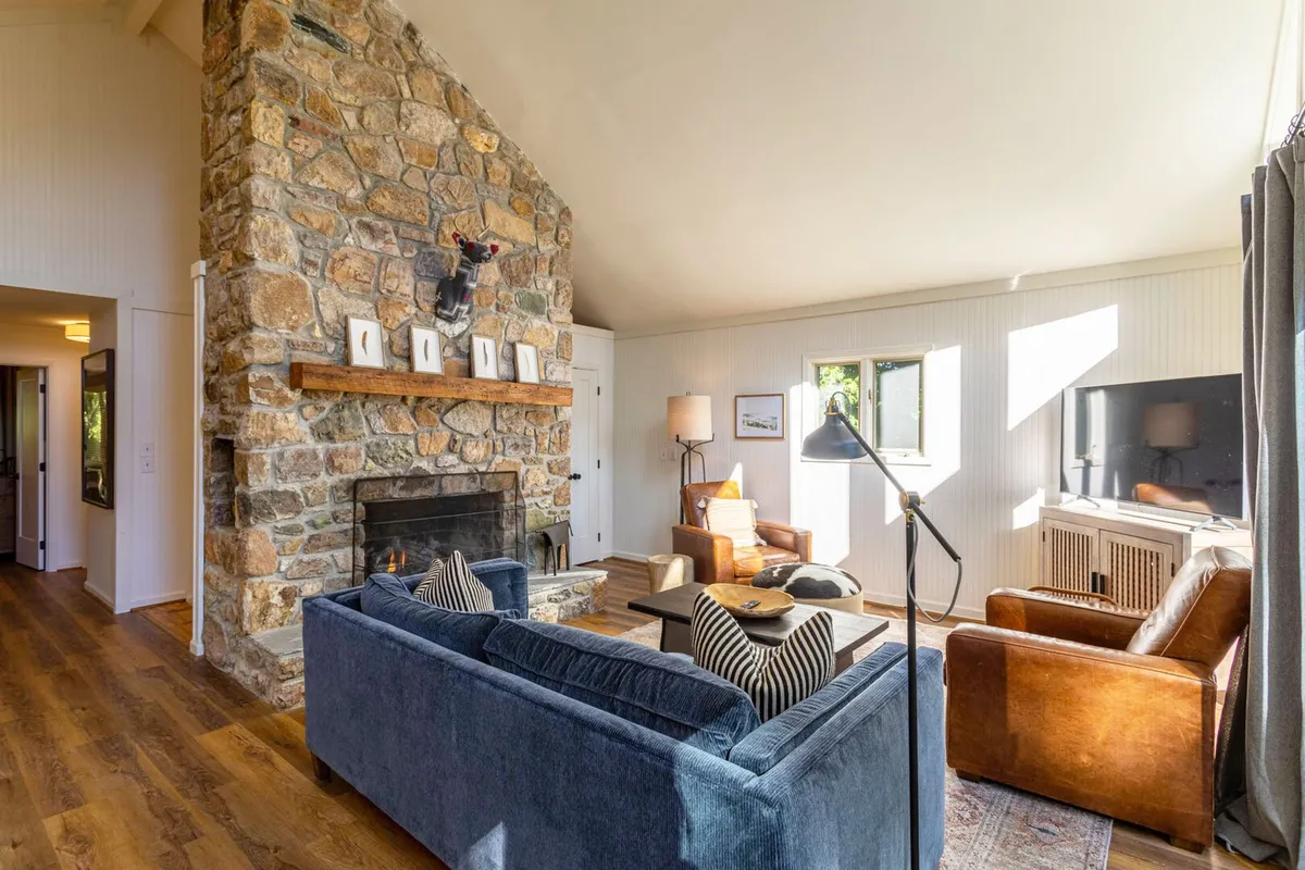 A cozy living area featuring a striking stone fireplace, comfortable seating arranged for relaxed gatherings, and expansive windows that let in abundant natural light and offer a glimpse of the outdoors. The warm tones and textures evoke comfort and seasonal retreat, perfect for unwinding after a day of mountain adventures.