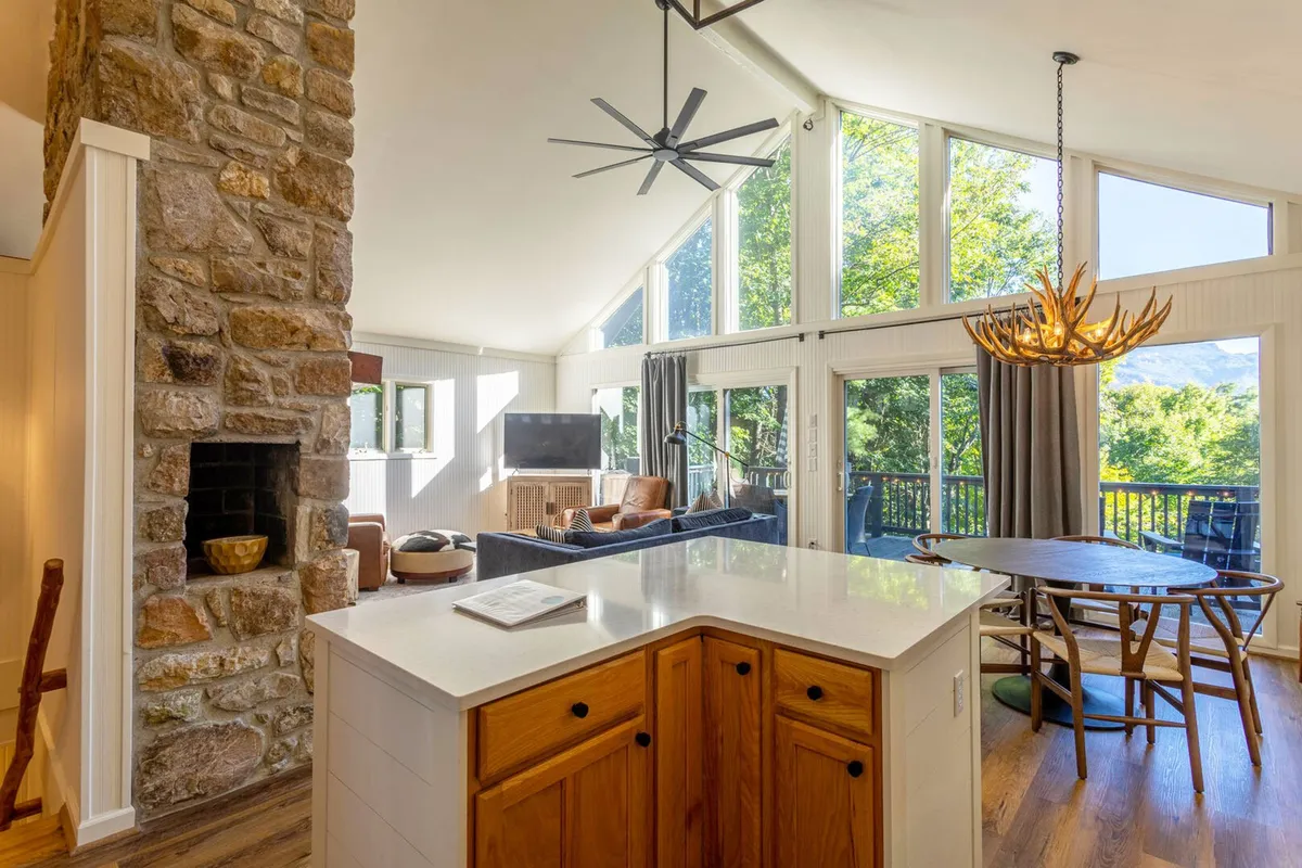 An expansive, open-concept interior highlighted by a stone fireplace, large windows framing beautiful views, a spacious dining table, modern chandelier, and comfortable seating. The space feels airy and light-filled, emphasizing both togetherness and the connection to the mountain scenery.