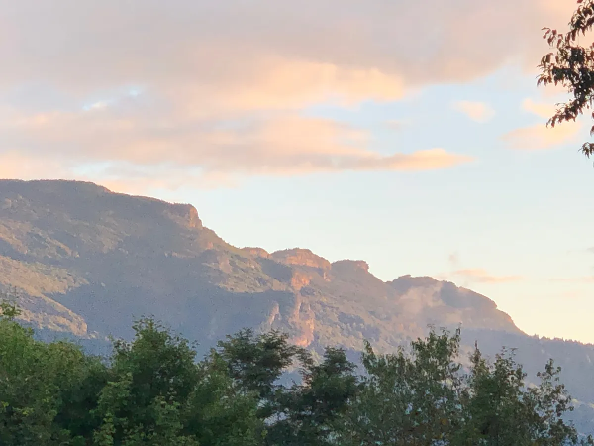 A sweeping view of a mountain range with dramatic clouds and lush foliage in the foreground. The scene is tranquil and invites outdoor adventure, with visible hiking trails snaking through the landscape. The vibrant colors hint at a sunset or sunrise, enhancing the sense of natural wonder.