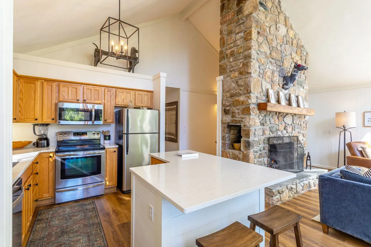 A warm and welcoming interior scene showcasing a stone fireplace, kitchen island, wood cabinetry, and a rustic chandelier. The space is clearly designed for cooking, entertaining, and gathering, embodying the spirit of group getaways and cozy evenings.