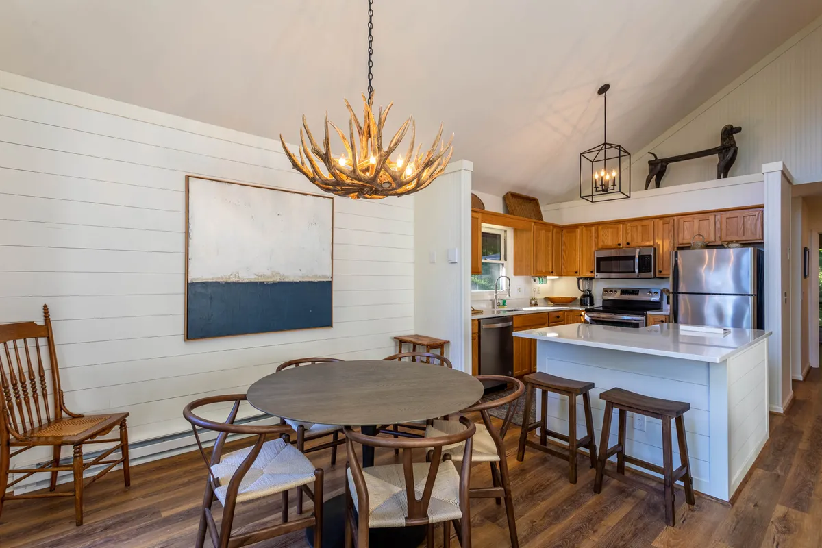 This image centers on a round dining table with wooden chairs beneath a striking antler chandelier, adjacent to the kitchen island. The table is set in a warmly lit environment, ideal for family meals and celebrations. The proximity to the kitchen is clear, visually connecting the two spaces.