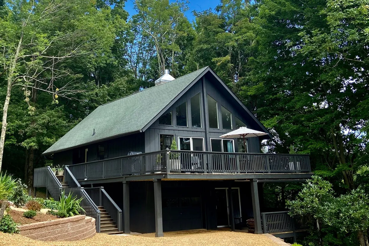 The exterior of the home featuring a large deck, a striking steep roofline, expansive windows, and lush greenery all around. The deck offers views into the surrounding forest, ideal for morning coffee or evening stargazing after a day of adventure.