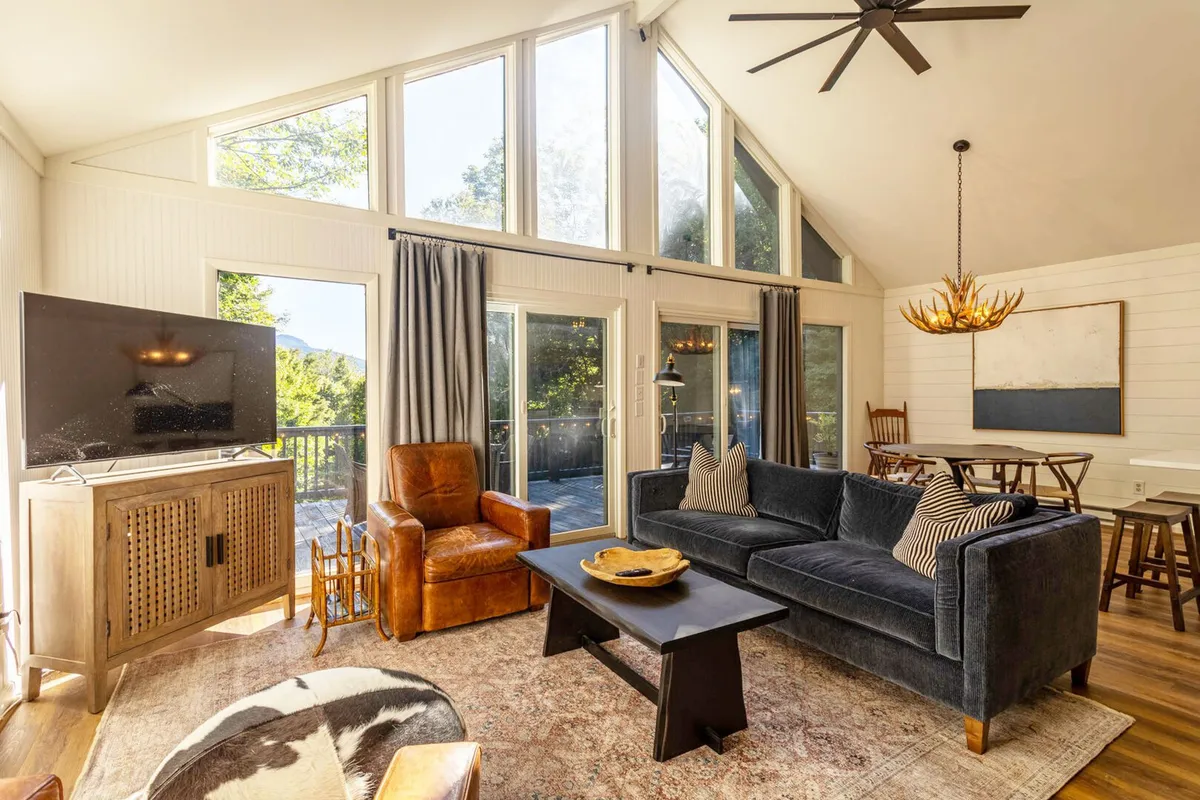 A spacious, inviting living room with a comfortable sofa, antique wooden chair, TV console, and a decorative chandelier. Large windows frame scenic views, creating a connection to the outdoors and providing a backdrop for relaxing, socializing, or watching a movie after a day of adventure.