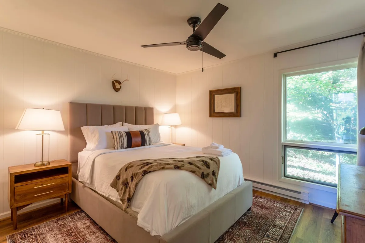 This image features a queen bed adorned with decorative pillows, a nightstand with a lamp, and a large window letting in ample natural light. The overall look is calm and inviting, suggesting restful mornings and peaceful evenings. Soft color tones and the outside view through the window enhance the room’s serenity.