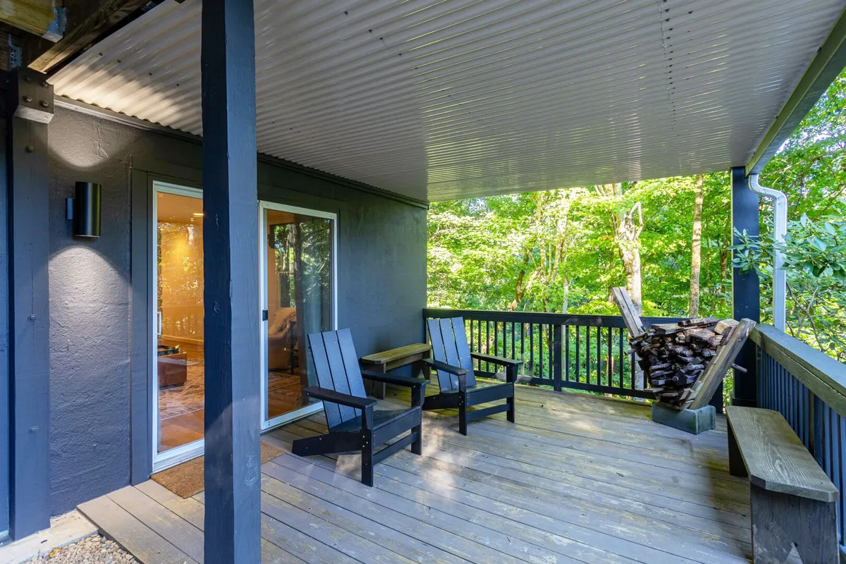 This exterior image features inviting wooden chairs arranged to face a lush view of trees, with a neat stack of firewood adding to the cozy, rustic ambiance. The scene suggests a perfect spot for relaxing, reading, or enjoying quiet moments surrounded by nature.