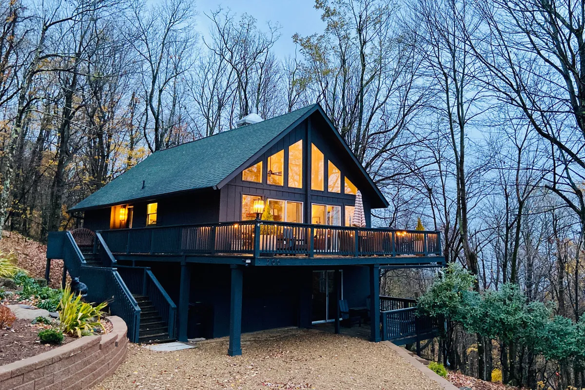The image shows the exterior of a cozy single-family home nestled among tall trees, with expansive windows and a spacious wooden deck overlooking the mountains. The scene suggests a tranquil escape with panoramic views, inviting guests to relax outdoors and savor the natural beauty.
