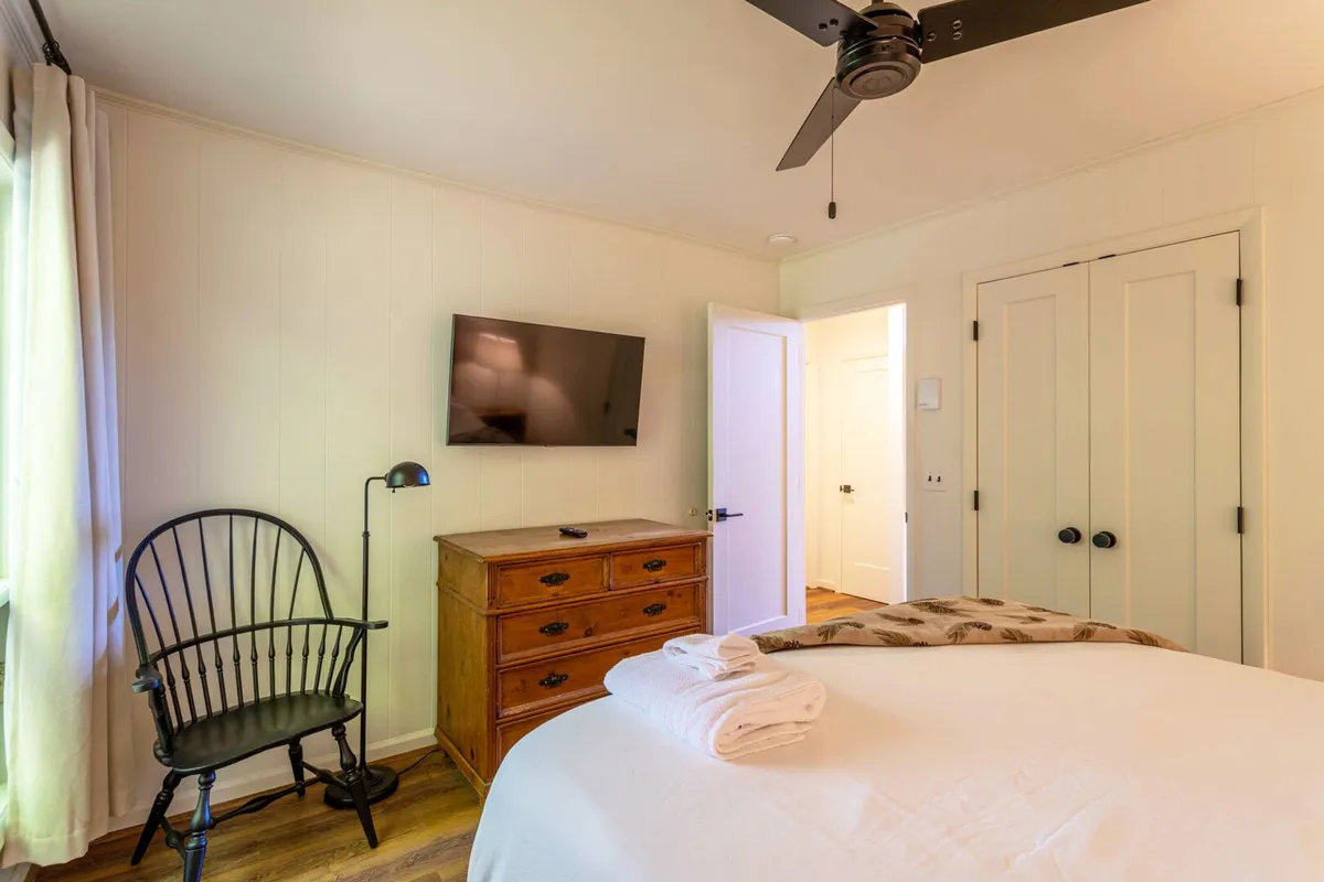 A queen bed topped with towels is the centerpiece, accompanied by a wooden dresser, a black chair, and a TV, all set in a relaxing atmosphere with morning light streaming in. The space feels ready for guests to unwind and recharge.