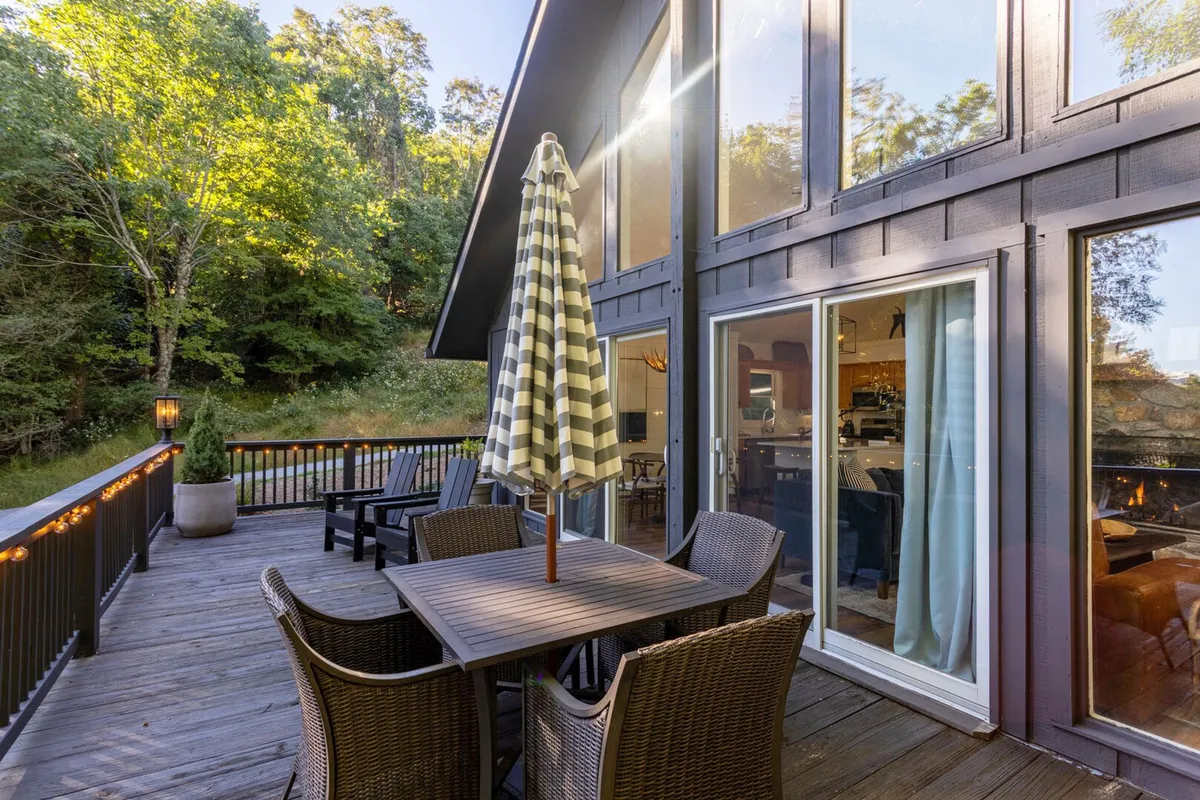 An inviting outdoor dining area with a striped umbrella, large windows reflecting the trees, and a backdrop of forested surroundings. The space feels perfect for al fresco family meals and soaking up the natural setting.